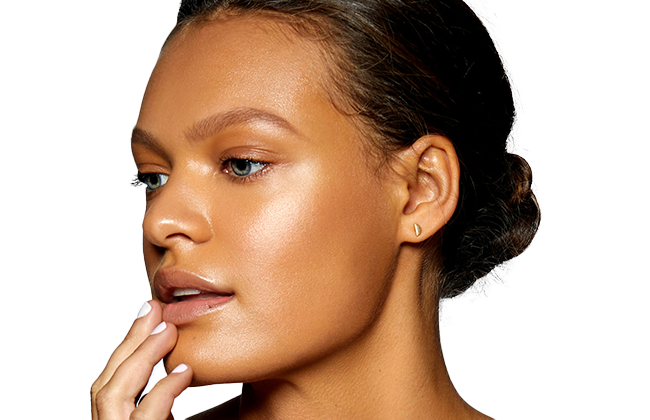 Close-up of woman with glowing bronzed skin, green eyes, low bun hairstyle, and small gold teardrop earring.